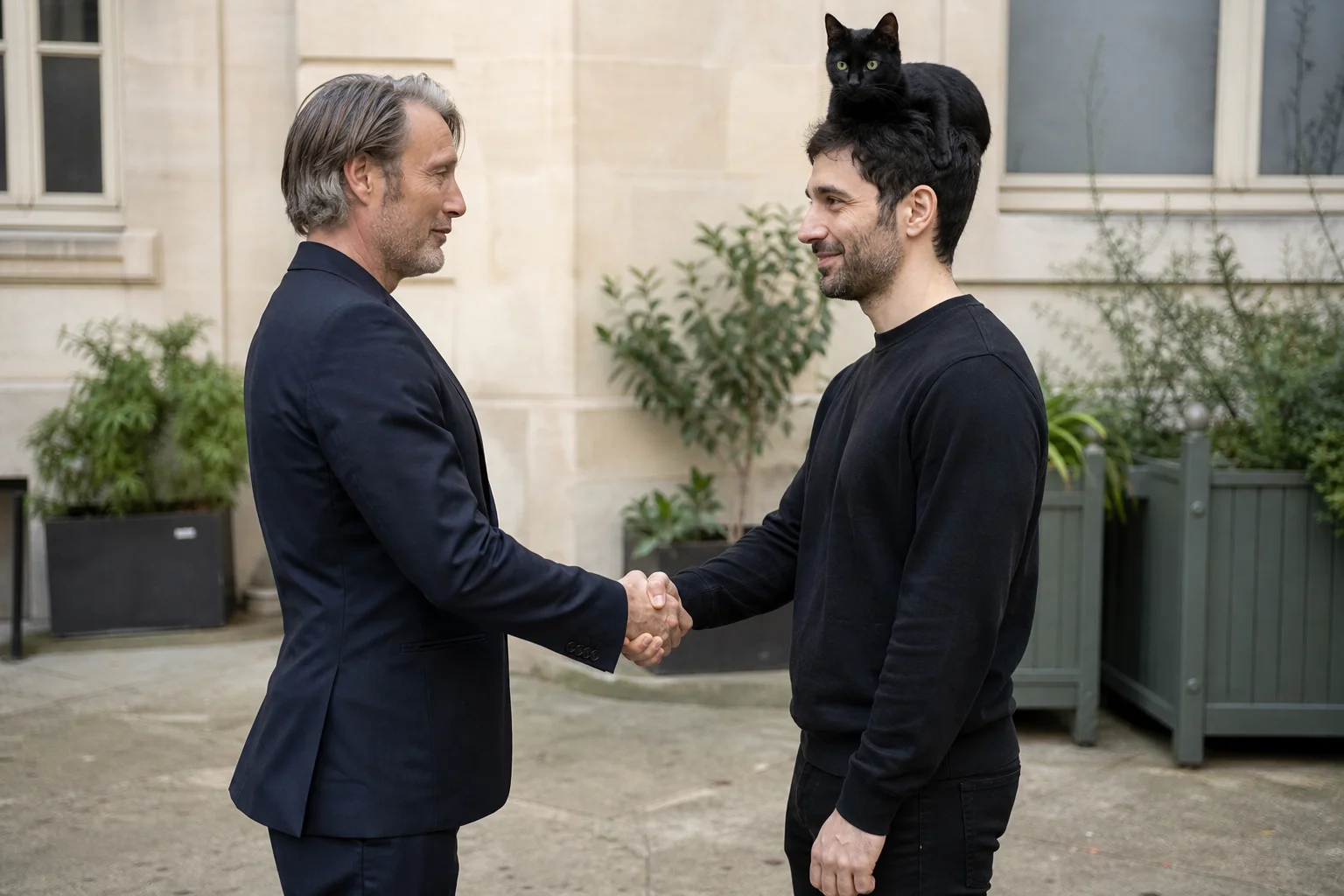 Two men warmly shake hands face-to-face outdoors while a black cat with vivid green eyes sits atop one man’s head in soft natural daylight.