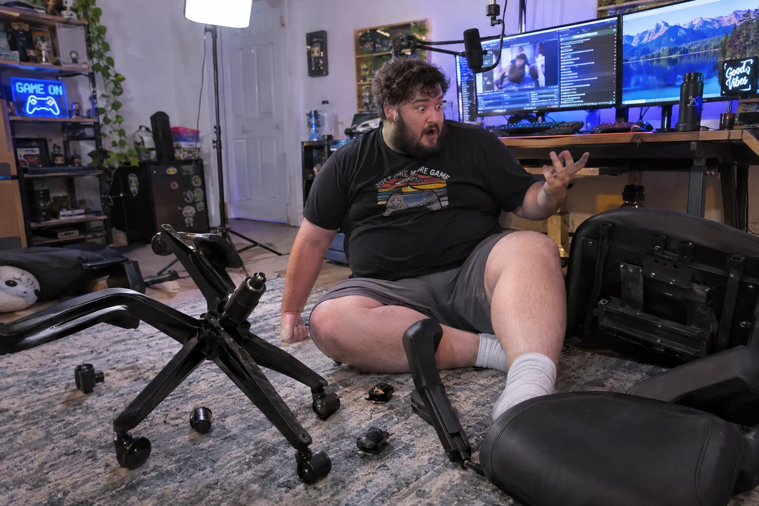 An overweight male streamer sits startled on the floor beside a broken rolling office chair in a cluttered home streaming room, surrounded by scattered chair parts, monitors, a microphone, and colorful studio lighting.